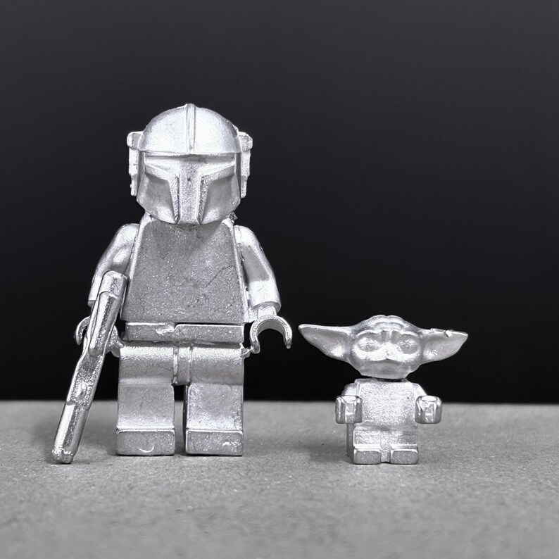 May include: Two silver-colored Lego figures: a Mandalorian with a helmet and blaster, and a small Grogu figure. The figures are on a gray surface with a black background. The Mandalorian figure is approximately 4 inches tall.