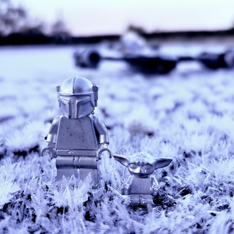 May include: Two silver-colored Lego figures of The Mandalorian and Grogu standing in a field of frosted grass. A blurred spaceship is in the background. The figures are detailed with helmets and armor.