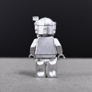 May include: A silver-colored minifigure with a helmet and rectangular torso. The figure has a detailed design, including a visor and a small antenna on top of the helmet. The figure is standing on a gray surface.