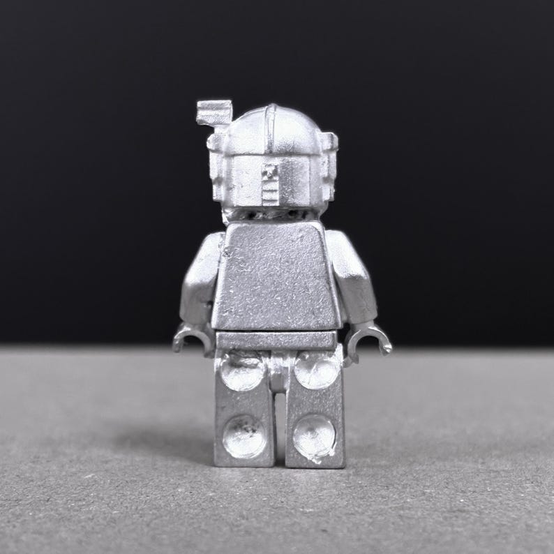 May include: A silver-colored minifigure with a helmet and rectangular torso. The figure has a detailed design, including a visor and a small antenna on top of the helmet. The figure is standing on a gray surface.