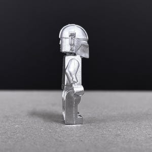 May include: A silver-colored minifigure with a helmet and armor. The figure is standing upright, with arms at its sides. The helmet has a distinctive shape, and the figure appears to be made of metal.