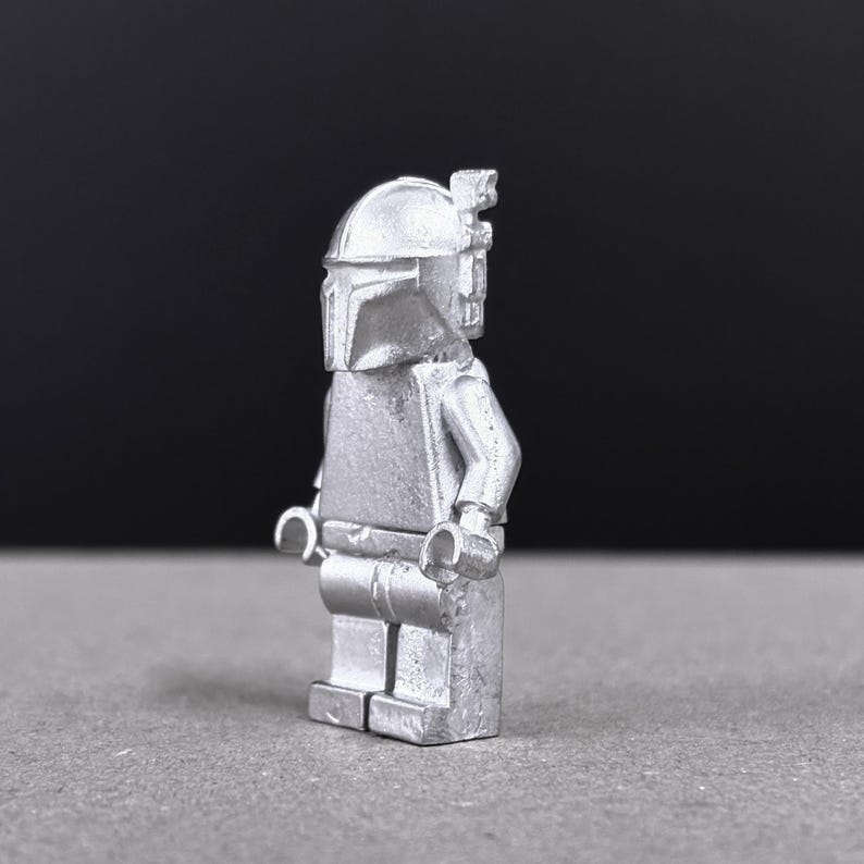 May include: A silver-colored Mandalorian figure, reminiscent of a popular science fiction character. The figure is a stylized block figure with a helmet, torso, arms, and legs. The figure is standing on a gray surface with a black background.