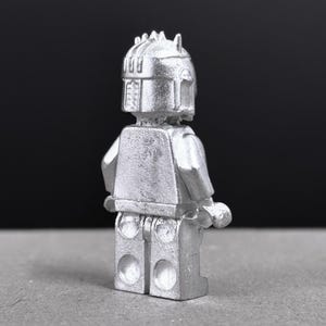 May include: A silver-colored figurine of a character with a helmet and armor, reminiscent of a space warrior. The figure has a blocky design, with a detailed helmet and a textured surface. The figure is standing upright.