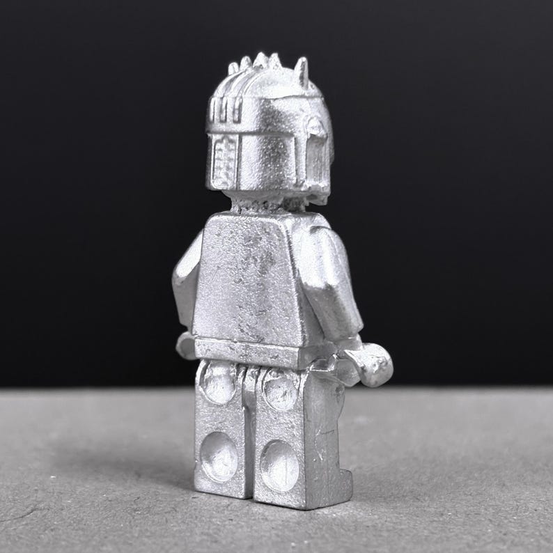 May include: A silver-colored figurine of a character with a helmet and armor, reminiscent of a space warrior. The figure has a blocky design, with a detailed helmet and a textured surface. The figure is standing upright.