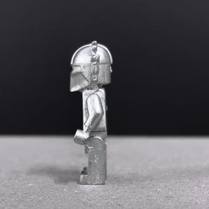May include: A silver-colored, stylized figurine of a character wearing a helmet and armor. The figure is standing upright with arms at its sides. The background is a gradient of gray and black.