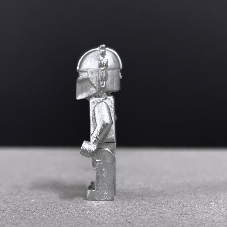 May include: A silver-colored, stylized figurine of a character wearing a helmet and armor. The figure is standing upright with arms at its sides. The background is a gradient of gray and black.