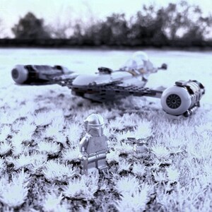 May include: A Lego model of a spaceship with two engines and a cockpit, along with two Lego figures, one resembling a Mandalorian and the other a small Grogu. The scene is set on a field of white, possibly frosted grass.