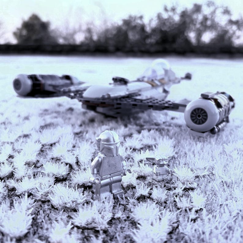 May include: A Lego model of a spaceship with two engines and a cockpit, along with two Lego figures, one resembling a Mandalorian and the other a small Grogu. The scene is set on a field of white, possibly frosted grass.