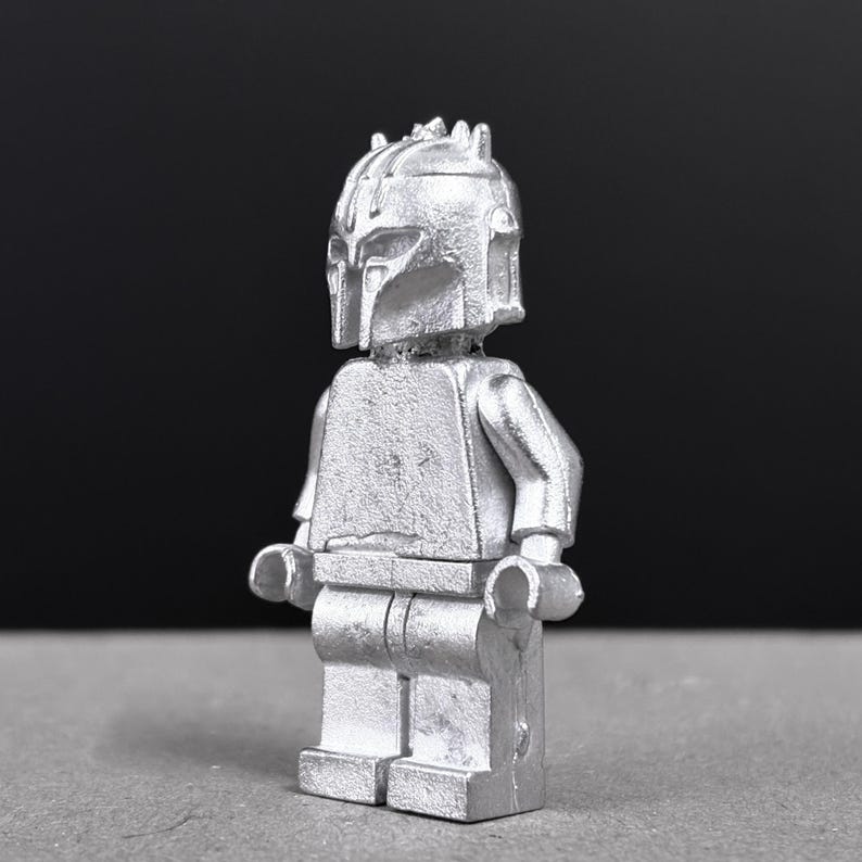 May include: A silver-colored minifigure with a helmet and armor, reminiscent of a Mandalorian character. The figure has a detailed helmet design and is posed against a dark background. The overall aesthetic is metallic and futuristic.