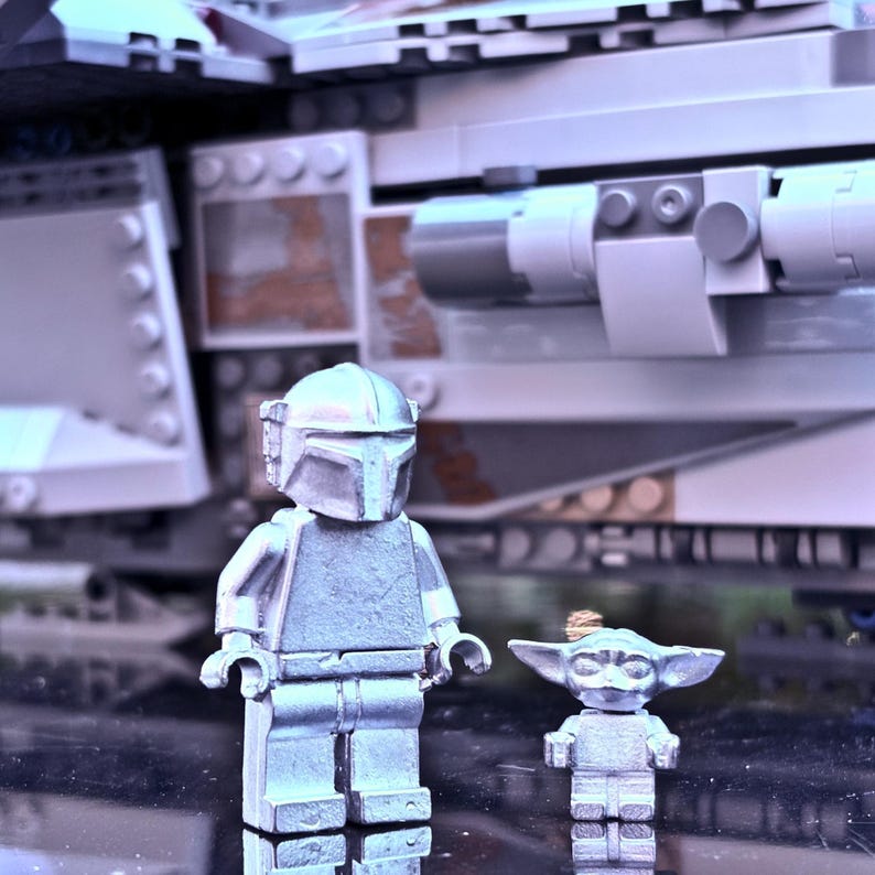 May include: Two silver-colored Lego figures of The Mandalorian and Grogu, standing in front of a Lego spaceship. The Mandalorian figure is larger and has a helmet. Grogu is smaller with large ears. The background is a light gray Lego structure.