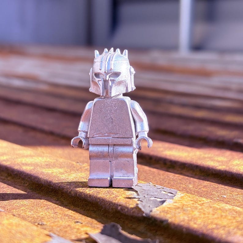 May include: A silver-colored Lego figure of a character with a helmet and armor. The figure is standing on a weathered, rusty metal surface. The figure's design is detailed, with visible lines and shapes.