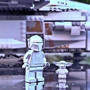 May include: A silver-colored Lego Mandalorian figure and a Lego Baby Yoda figure stand in front of a Lego spaceship. The Mandalorian figure is wearing a helmet and armor. The Baby Yoda figure has large ears and a small body. The spaceship is gray and white.