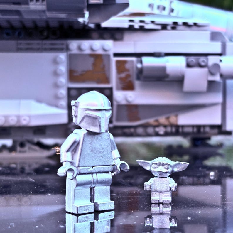 May include: A silver-colored Lego Mandalorian figure and a Lego Baby Yoda figure stand in front of a Lego spaceship. The Mandalorian figure is wearing a helmet and armor. The Baby Yoda figure has large ears and a small body. The spaceship is gray and white.