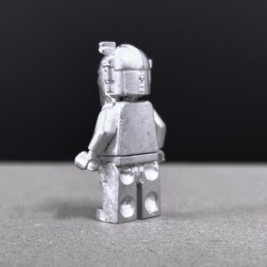 May include: A silver-colored figurine of a minifigure, viewed from the back. The figure has a helmet and a rectangular torso with arms and legs. The background is a dark gray.