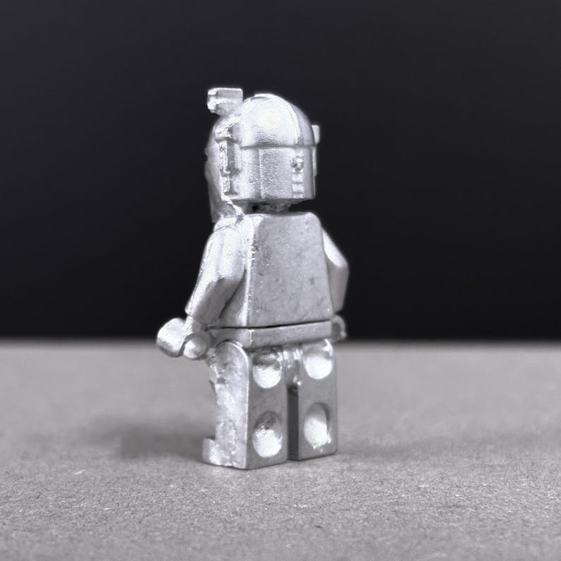 May include: A silver-colored figurine of a minifigure, viewed from the back. The figure has a helmet and a rectangular torso with arms and legs. The background is a dark gray.