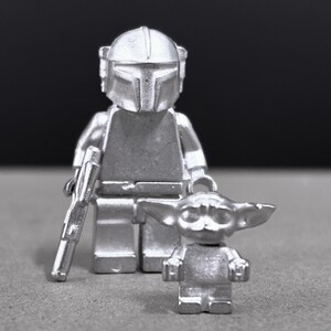 May include: Silver-colored Mandalorian and Grogu Lego-style figures. The Mandalorian figure holds a blaster. The Grogu figure is smaller and stands next to the Mandalorian. The figures are on a gray surface with a dark background.