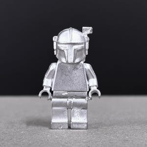 May include: A silver-colored Lego figurine of a Mandalorian character, featuring a helmet with a T-shaped visor and a rectangular antenna. The figure has articulated arms and legs, and is set against a dark background.
