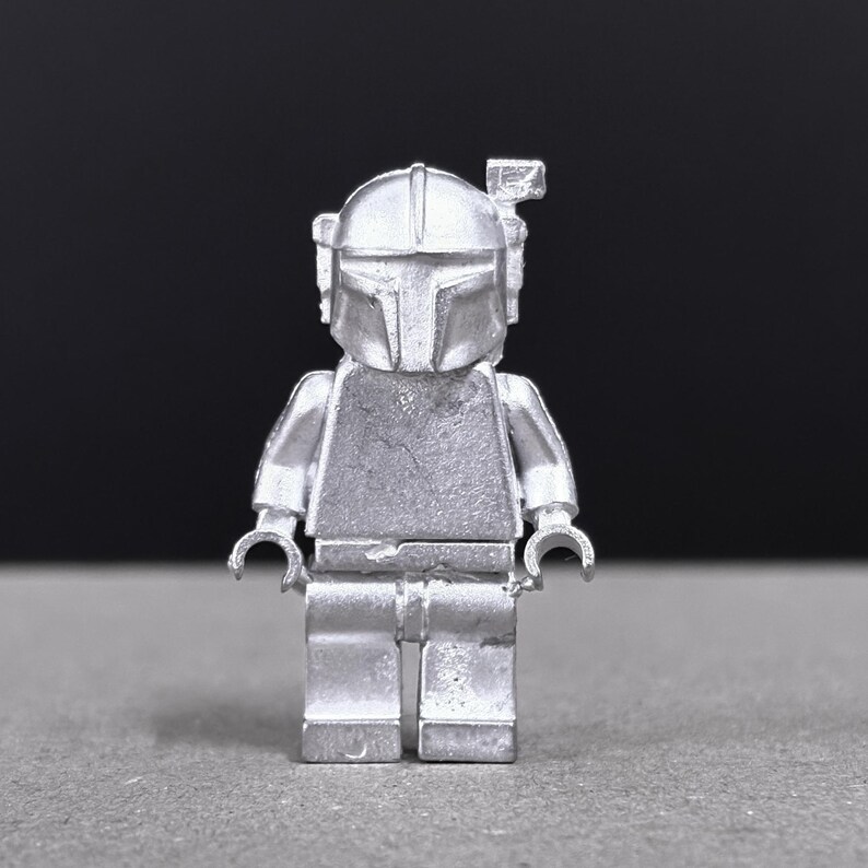 May include: A silver-colored Lego figurine of a Mandalorian character, featuring a helmet with a T-shaped visor and a rectangular antenna. The figure has articulated arms and legs, and is set against a dark background.