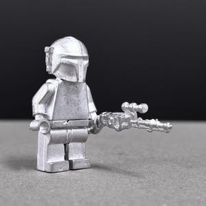 May include: A silver-colored Mandalorian figure, a minifigure, holding a blaster gun. The figure has a helmet and armor, and is posed against a dark background. The toy is a collectible item.