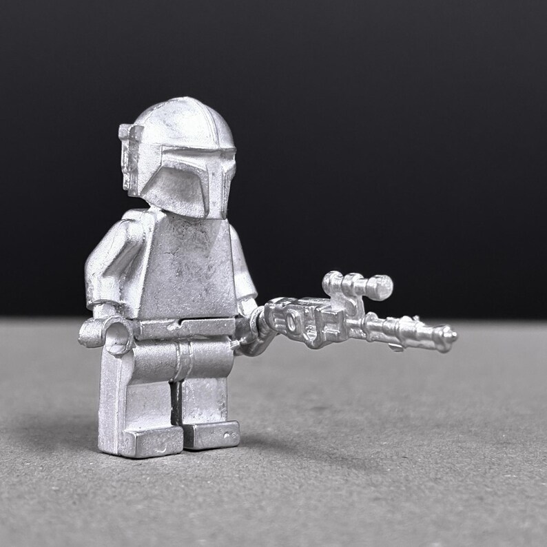 May include: A silver-colored Mandalorian figure, a minifigure, holding a blaster gun. The figure has a helmet and armor, and is posed against a dark background. The toy is a collectible item.