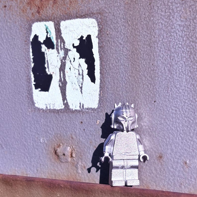 May include: A silver-colored Lego minifigure with a helmet and armor stands in front of a weathered, textured surface. The background features a faded white rectangle with black shapes.