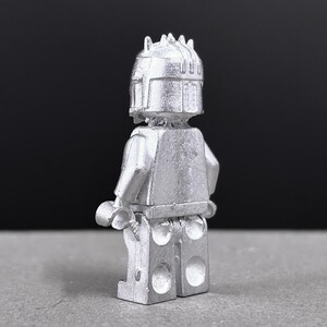 May include: A silver-colored Lego figure, viewed from the back, with a detailed helmet and armor. The figure has a classic Lego design with articulated arms and legs, and a textured surface. The background is a dark gray.
