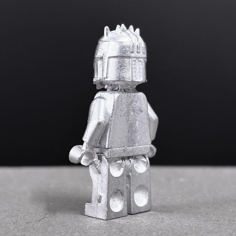 May include: A silver-colored Lego figure, viewed from the back, with a detailed helmet and armor. The figure has a classic Lego design with articulated arms and legs, and a textured surface. The background is a dark gray.