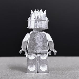 May include: A silver-colored Lego minifigure, viewed from the back, with a detailed helmet and crown. The figure has a blocky torso, arms, and legs, with a textured surface. The background is a solid dark gray.