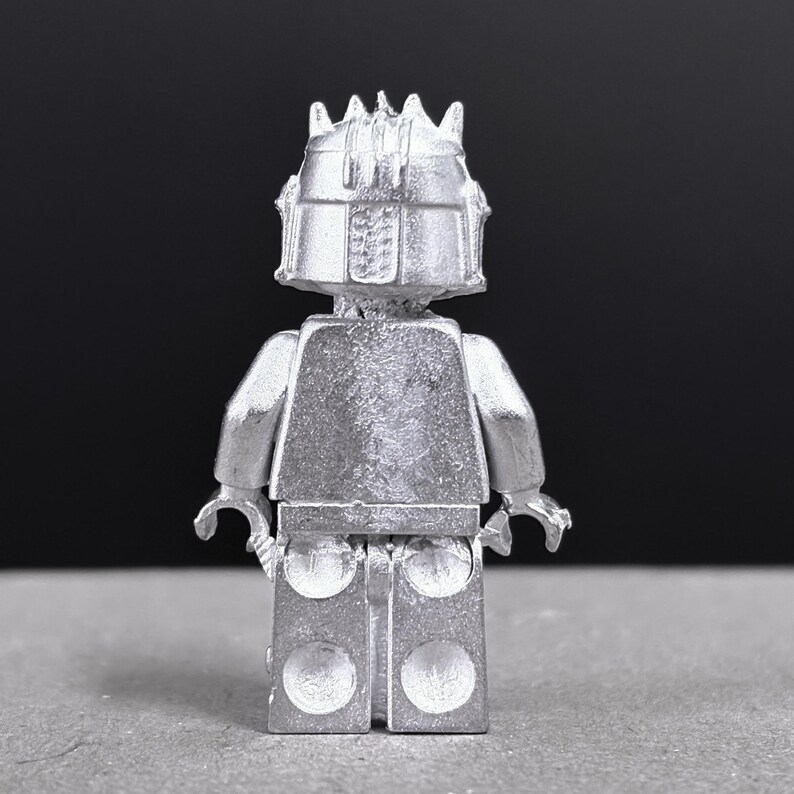 May include: A silver-colored Lego minifigure, viewed from the back, with a detailed helmet and crown. The figure has a blocky torso, arms, and legs, with a textured surface. The background is a solid dark gray.