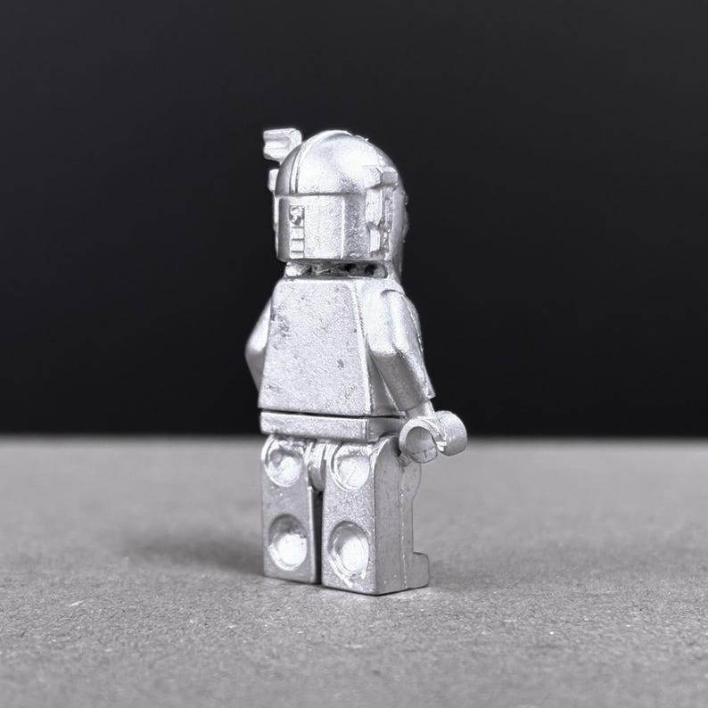 May include: A silver-colored figurine of a character with a helmet and rectangular torso. The figure has a classic blocky design, with visible details on the helmet and body. The figure is standing on a gray surface.