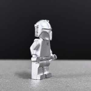 May include: A silver-colored Mandalorian figure, a collectible toy, stands in profile against a dark background. The figure has a helmet, armor, and a weapon. The toy is made of a solid material and has a smooth, metallic finish.