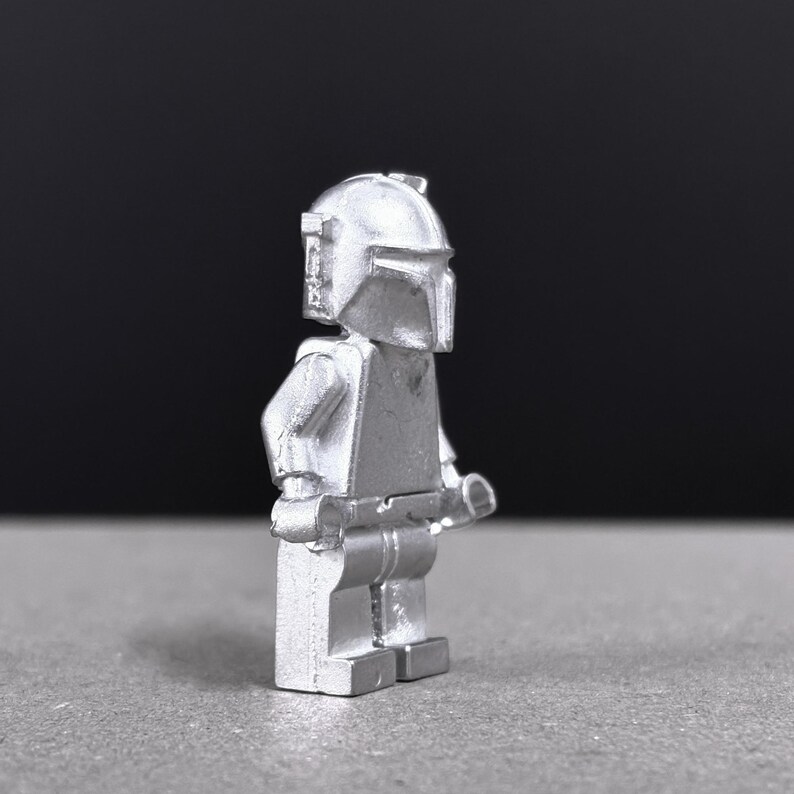 May include: A silver-colored Mandalorian figure, a collectible toy, stands in profile against a dark background. The figure has a helmet, armor, and a weapon. The toy is made of a solid material and has a smooth, metallic finish.