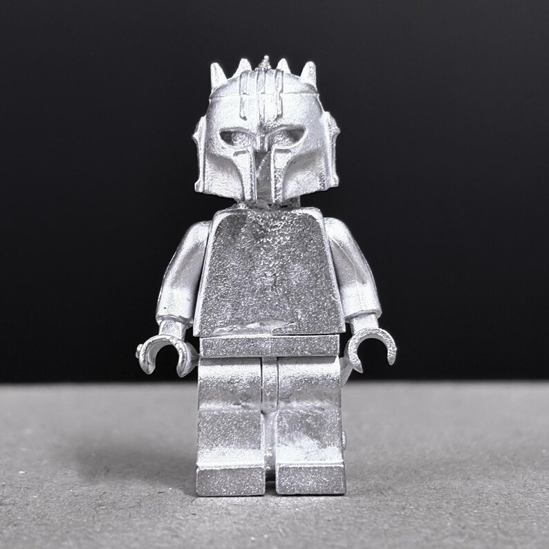 May include: A silver-colored Lego minifigure with a helmet and armor. The figure has a detailed design, including a crown-like structure on the helmet. The background is a solid black, highlighting the metallic sheen of the toy.