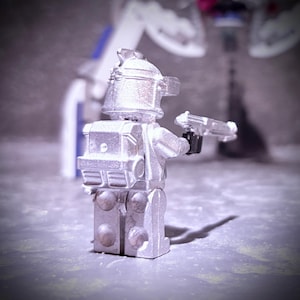 May include: A silver-colored Lego minifigure with a helmet, backpack, and a transparent gun. The figure is standing on a gray surface, with a blurred background featuring a white and blue structure.