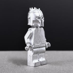 May include: A silver-colored Lego minifigure with a helmet and armor. The figure is standing upright with arms at its sides. The helmet has a distinctive crest and visor. The background is a dark gray.