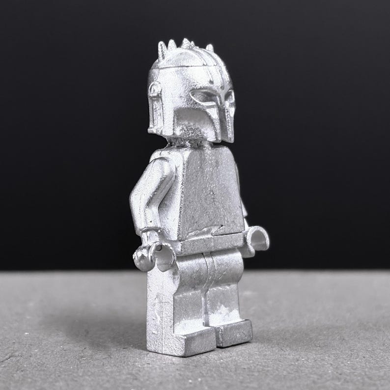 May include: A silver-colored Lego minifigure with a helmet and armor. The figure is standing upright with arms at its sides. The helmet has a distinctive crest and visor. The background is a dark gray.
