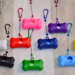 Könnte beinhalten: Eine Auswahl an farbenfrohen Hundekotbeutelspendern in Weiß, Schwarz, Rot, Pink, Grün, Blau, Lila und Gelb. Jeder knochenförmige Spender hat einen Metallclip zur Befestigung.