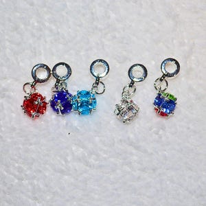 May include: A collection of five colorful charm pendants. Each pendant features a spherical, faceted gemstone in red, blue, light blue, clear, and a multi-colored combination. The charms are attached to silver-colored metal loops.