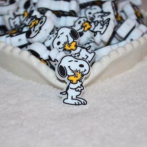 May include: White and black Snoopy-themed charms in a white, scalloped-edge dish. The charms feature Snoopy and Woodstock. The charms are likely for jewelry making or other crafts.