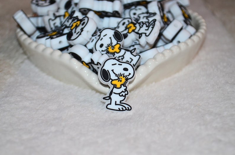 Pode incluir: Berloques brancos e pretos com tema Snoopy em um prato branco com borda ondulada. Os berloques apresentam Snoopy e Woodstock. Os berloques s&atilde;o provavelmente para fazer joias ou outros artesanatos.