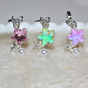 May include: Three star-shaped earrings in pink, green, and purple. Each earring features a larger, faceted star, a smaller silver star, and a white pearl. The earrings are displayed on a white, textured surface.