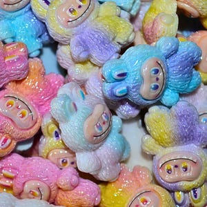 May include: A close-up of a pile of colorful, smiling monkey-like figurines. The figurines are in various pastel colors, including pink, blue, yellow, and purple. Each has a smiling face with large eyes and a small mouth. The overall theme is whimsical and playful.