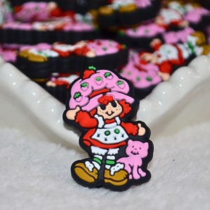 May include: A colourful charm featuring a cartoon character with red hair, a red dress, and a pink hat with strawberries. The charm is holding a pink cat and has a black border. Other similar charms are in the background.