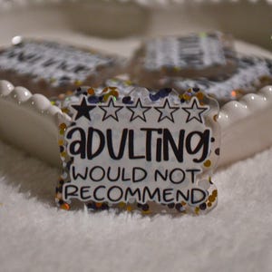 Puede incluir: Insignia de acrílico transparente con el texto "ADULTING: WOULD NOT RECOMMEND" en letras negras, acentuado con purpurina dorada, azul y naranja. La insignia presenta un diseño de estrellas y está sobre un fondo blanco.