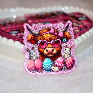 May include: A pink glittery Easter decoration featuring a Highland cow wearing heart-shaped sunglasses and a pink bow. Two bunnies and decorated Easter eggs are also featured. The background is a white heart-shaped dish.
