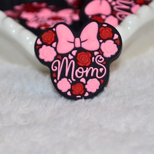 May include: A black shoe charm in the shape of a mouse head with a pink bow and the word "Mom". The charm is decorated with pink and red roses. A few other charms are visible in the background.