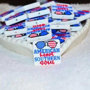 May include: White rectangular beads with a patriotic theme, featuring sunglasses with red, white, and blue designs. The text "AMERICAN heart SOUTHERN soul" is displayed in blue and red. The beads are in a white, heart-shaped dish.