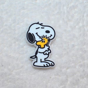 Pode incluir: Um pingente branco de Snoopy com contornos pretos, segurando um p&aacute;ssaro amarelo. O pingente &eacute; um personagem de desenho animado da hist&oacute;ria em quadrinhos Peanuts. O pingente est&aacute; em uma superf&iacute;cie branca texturizada.