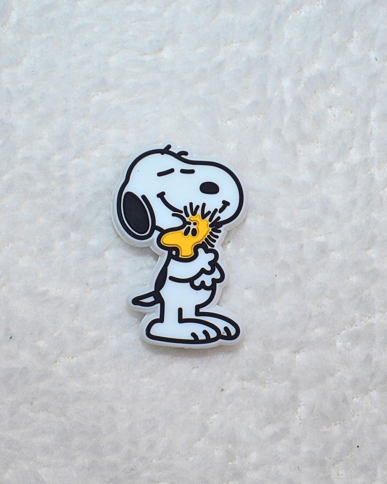 Pode incluir: Um pingente branco de Snoopy com contornos pretos, segurando um p&aacute;ssaro amarelo. O pingente &eacute; um personagem de desenho animado da hist&oacute;ria em quadrinhos Peanuts. O pingente est&aacute; em uma superf&iacute;cie branca texturizada.