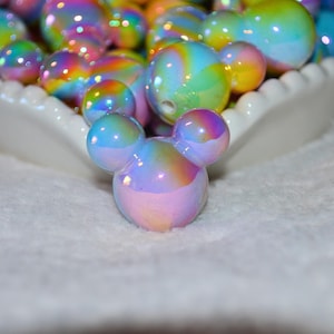 May include: Iridescent Mickey Mouse-shaped beads in a white scalloped dish. The beads are a mix of colours, including pink, yellow, blue, and green. The beads are on a white, textured surface.